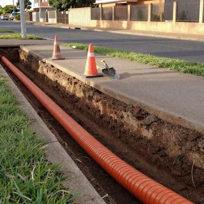 An open pavement trench with orange fibre duct being laid, cones and a worker alongside